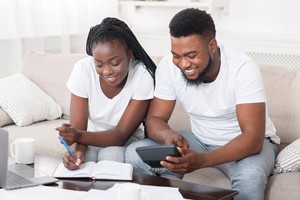 Young happy african american couple planning family budget together, sitting on sofa at home, using calculator and taking notes about contraceptives and birth control
